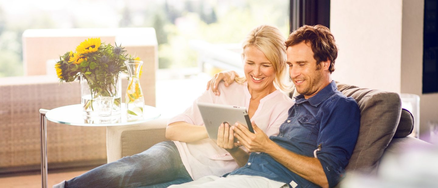 Mann und Frau sitzen auf der Couch. Frau zeigt Mann etwas auf dem iPad __ Man and woman are sitting on the couch. Woman is showing sth. on the iPad