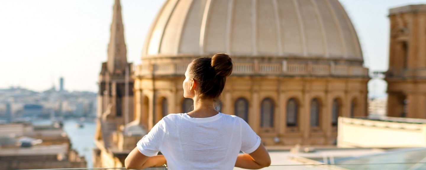 A brunette woman stands on a balcony at a height against the background of the city and the church with a dome. Focus on the woman. Back view